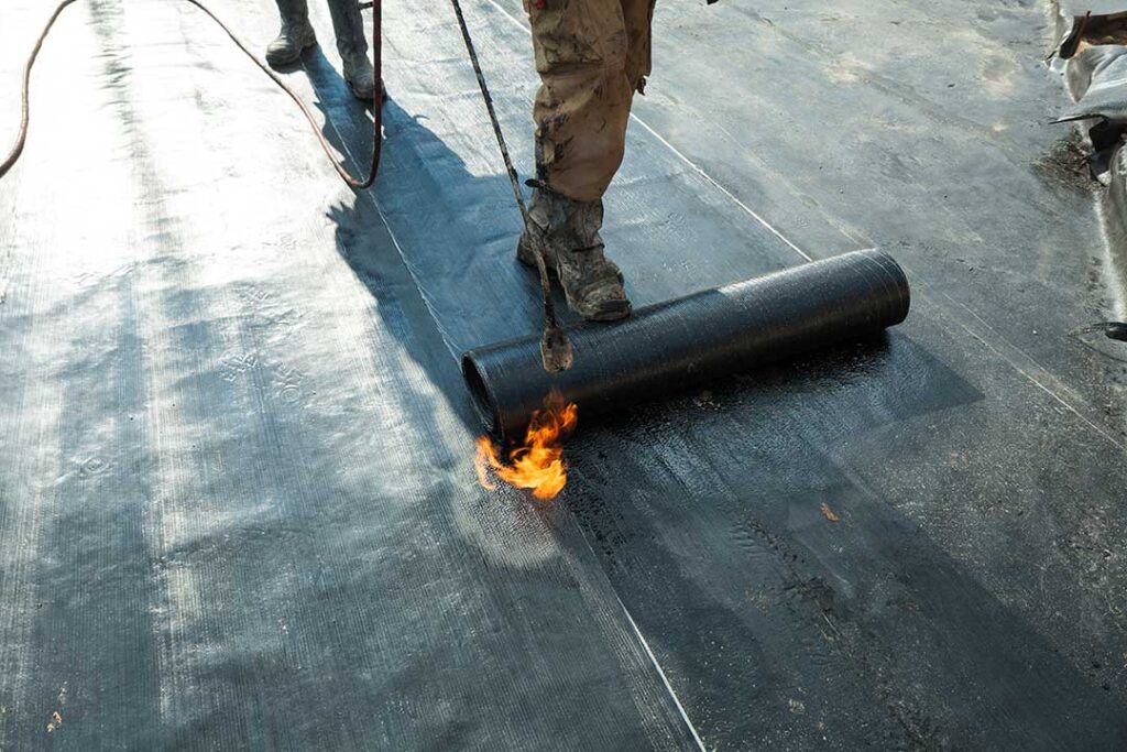 A roofer examines metal flashing and a rubber vent collar around a pipe on a shingled roof.