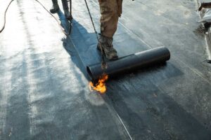 A roofer examines metal flashing and a rubber vent collar around a pipe on a shingled roof.